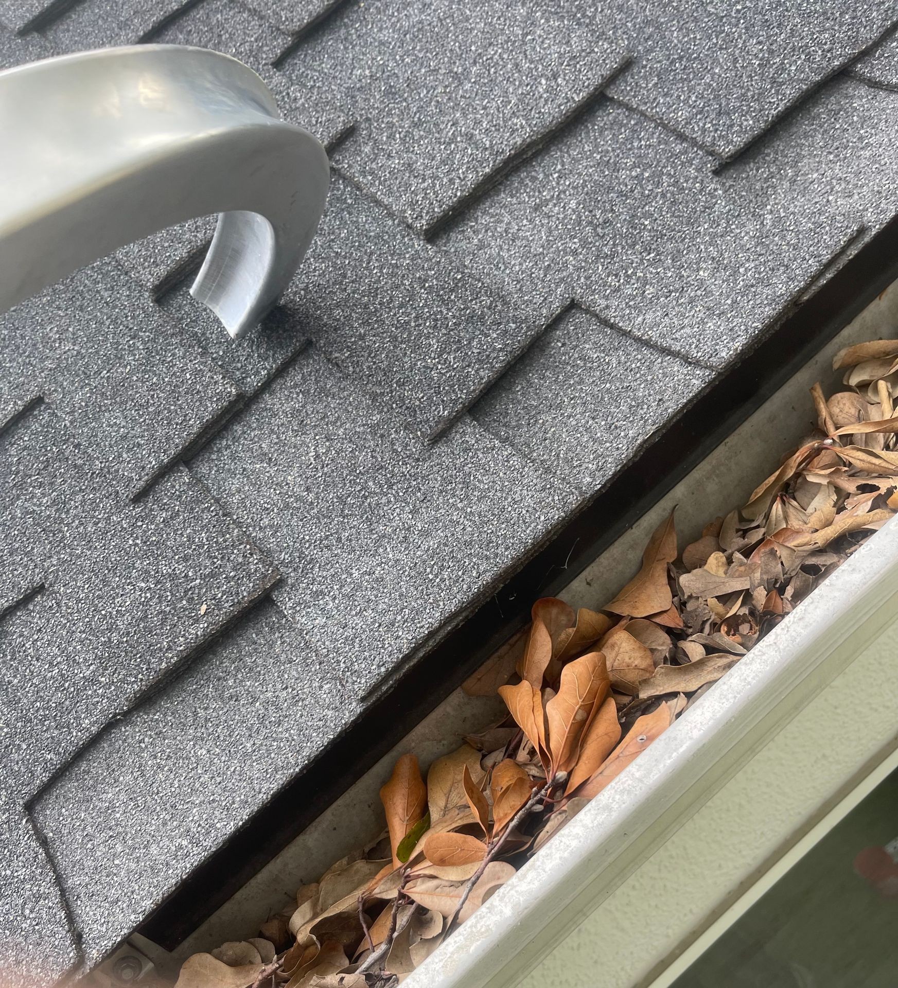 Roof shingles and a gutter filled with dry leaves, with a metal tool placed on the roof.