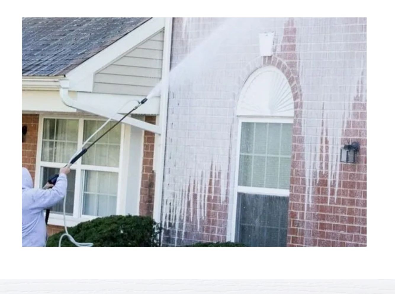 Person pressure-washing the exterior of a house, cleaning the brick wall and windows.